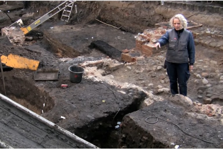 Vi sætter vore spor på toppen af historiske lag.</br>Udgravning ved Bispetorv 2009, Aarhus. Arkæologen står på fundamentet af et Middelalder kanikkehus i munkesten og peger på - muligvis - et brøndudløb.