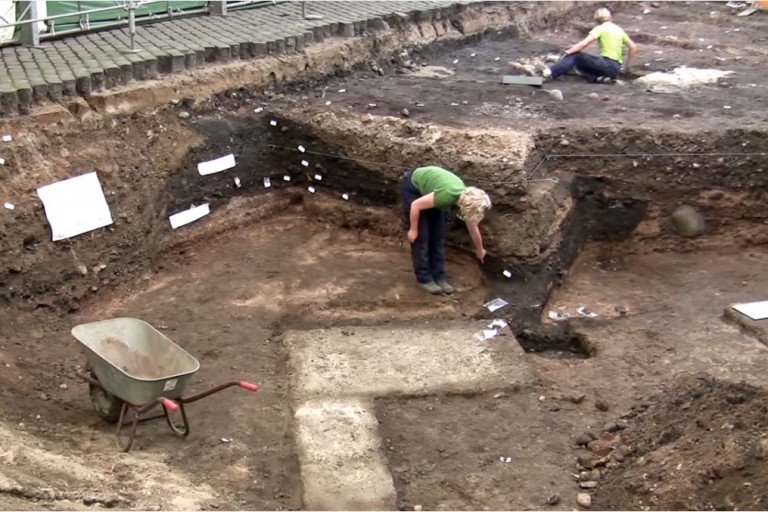 Vi sætter vore spor på toppen af historiske lag.</br>Udgravning ved Bispetorv 2009, Aarhus, Beton-elementet er resterne af en bunker fra 2. verdenskrig, Det mørke område, som arkæologen peger på, er et vikinge grubehus.